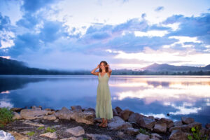 Beautiful high school senior in long flowing dress poses by the lake as the sunset and sky reflect like cotton candy on the water.